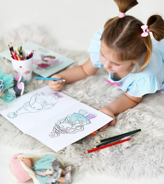 Küçük kız evde yumuşak halıya uzanmış renkli kalemlerle bebeklerin resimlerini çiziyor. Üst görünüm — Stok fotoğraf