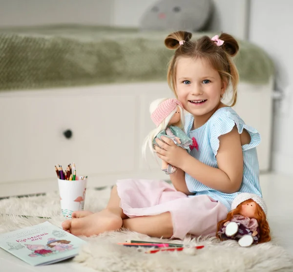 Милая улыбающаяся девочка сидит дома на мягком ковре, обнимаясь с куклами и рисуя картинки с красочными карандашами. — стоковое фото