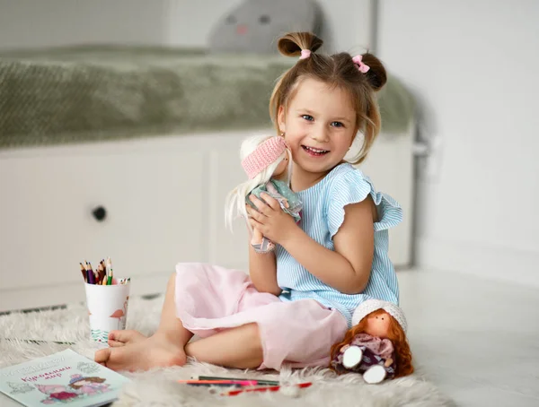 Счастливая девочка в домашнем платье сидит на мягком ковре дома обнимаясь играя с куклами и рисуя с красочными карандашами — стоковое фото