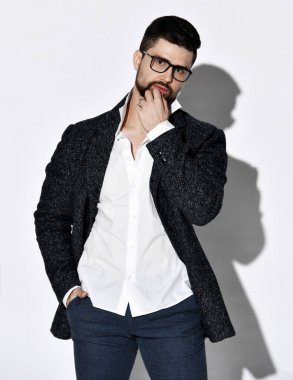 Düşünceli, şık sakallı, gözlüklü, tüvit ceketli ve kot pantolonlu adam cebinde ve çenesinde el ele duruyor.