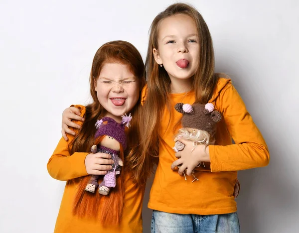 Портрет двух веселых подружек в оранжевых рубашках, стоящих вместе с куклами, обнимающими торчащими из своих языков — стоковое фото