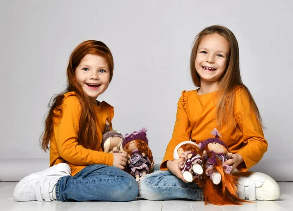 Dois felizes garotas risos amigos em camisolas laranja e jeans azuis sentam-se no chão brincando com suas bonecas felizes — Fotografia de Stock