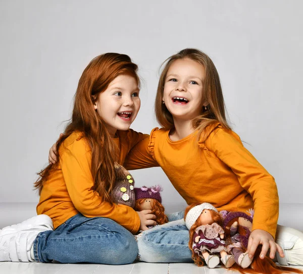 Two frolic laughing kid girls friends in similar clothes sit on floor in kindergarten playing with their dolls — Stock Photo, Image