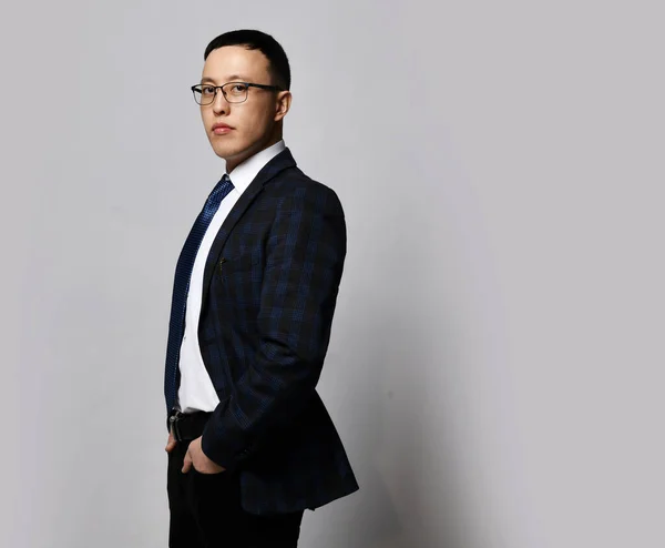 Young confident asian businessman in official suit, tie and glasses standing side to camera with copy space behind — Stock Photo, Image