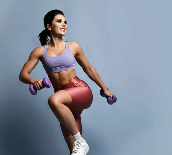 Joven hermosa morena fitness mujer en ropa deportiva haciendo ejercicio con mancuerna durante el entrenamiento deportivo sobre fondo púrpura — Foto de Stock