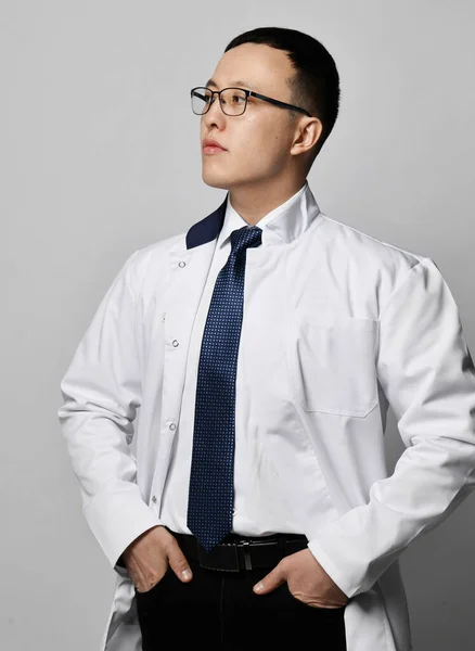 Potret dokter muda berkacamata, seragam gaun medis, kemeja putih dan dasi berdiri dengan tangan di saku — Stok Foto