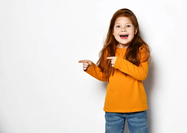Возбужденная рыжеволосая девочка в оранжевой толстовке стоит с открытым ртом в удивлении и указывает на пространство для копирования — стоковое фото
