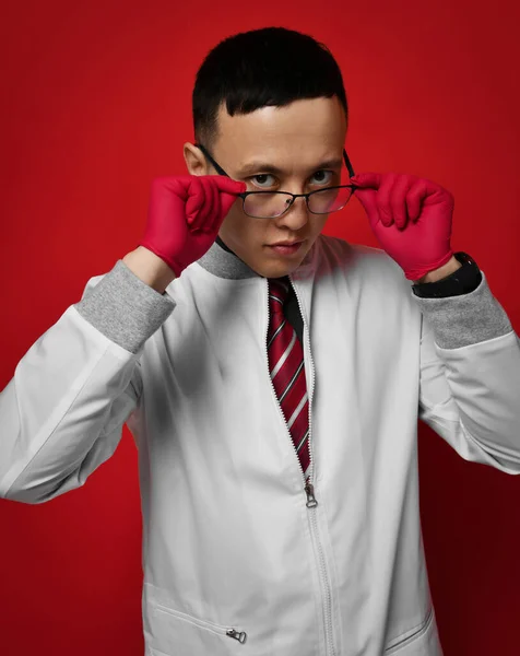 Portrait of young man doctor urologist or proctologist in medical gown and gloves putting on taking off his glasses — Stock Photo, Image