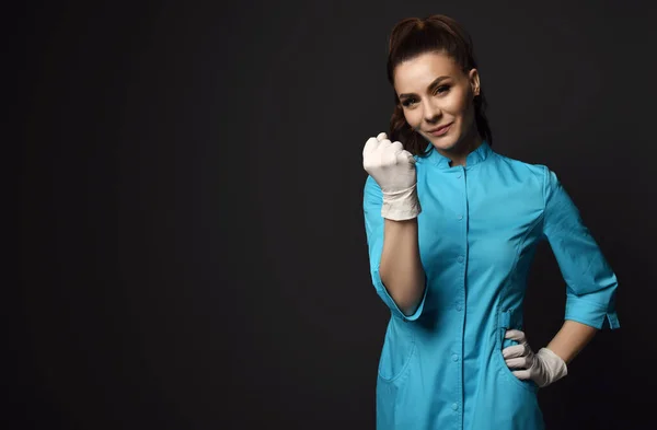 Молода усміхнена брюнетка лікар медсестра в блакитній медичній сукні і латексні рукавички потискають кулак на нас в копіювальному просторі — стокове фото