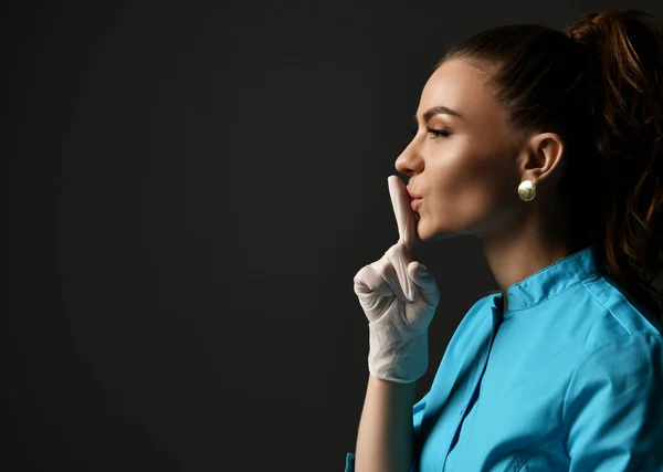 Young woman doctor in blue medical gown and latex gloves holds finger at her lips gesturing shh sign calling for silence — Stock Photo, Image