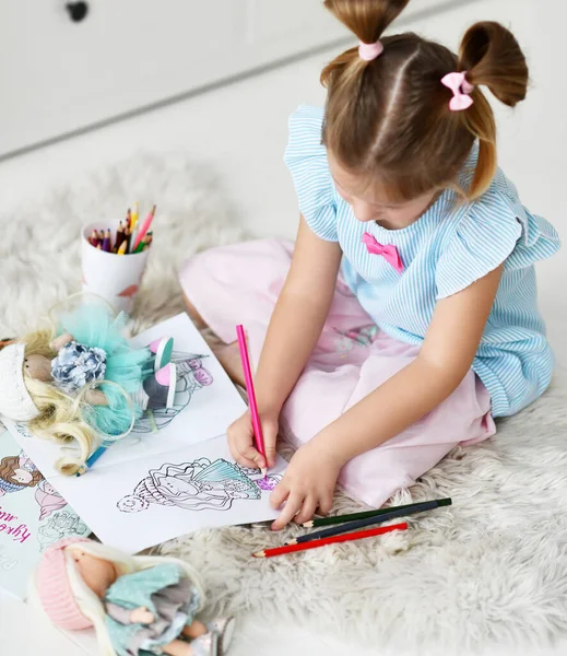 Menina criança com pães engraçados senta-se no tapete macio em casa desenho colorir fotos de bonecas com lápis coloridos. Vista superior — Fotografia de Stock