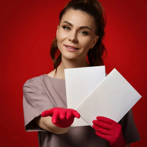 Giovane donna sorridente medico terapeuta in guanti protettivi in lattice in possesso di due fogli di carta spazi vuoti, indicando e guardando la fotocamera — Foto Stock