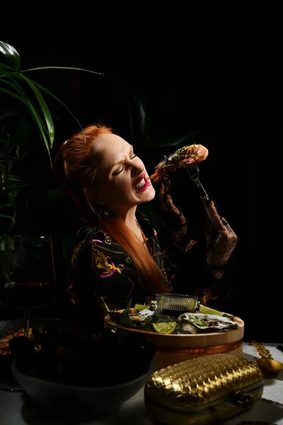 Rik rödhårig kvinna äter i skaldjur restaurang innehar en gaffel räkor eller havskräfta. Matbord som serverar färska ostron — Stockfoto
