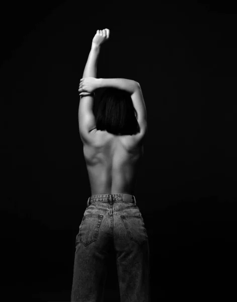 Mujer sexy está de pie en topless y en jeans de nuevo a la cámara sosteniendo la mano hacia arriba mostrando su forma de cuerpo perfecto. Blanco y negro —  Fotos de Stock