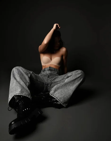 Sexig brunett kvinna bär pärla halsband, behå, breda jeans och massiva stövlar sitter på golvet gömmer ansiktet tittar åt sidan — Stockfoto