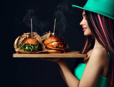 Güzel bir kadın aç ağızlı iki büyük burger sandviçi ve yeşil şapkalı buhar dumanı ile Blac 'a gülümsüyor.
