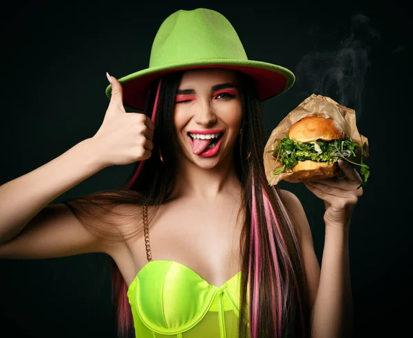 Mooie vrouw grote vegetarische rucola salade hamburger sandwich met stoom rook tonen duimen omhoog in groene hoed gelukkig glimlachen op zwart — Stockfoto