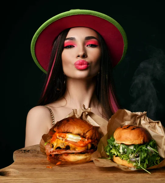 Mulher bonita segurar duas sanduíches de hambúrguer grande ofício com fome boca e fumaça de vapor em chapéu verde feliz sorrindo em blac — Fotografia de Stock