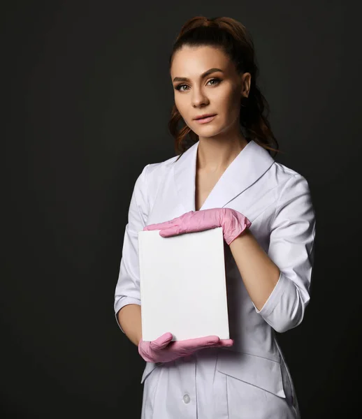 Joven bonita morena doctora terapeuta enfermera en uniforme blanco y guantes protectores sosteniendo hoja de papel en blanco — Foto de Stock