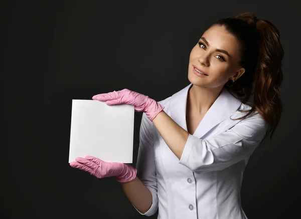 Fiatal mosolygós barna nő orvos terapeuta nővér fehér egyenruhában és védőkesztyű kezében üres papírlap — Stock Fotó