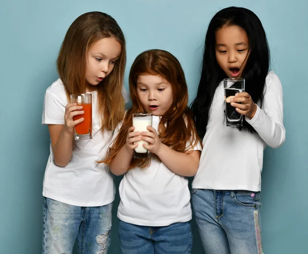 Three surprised socked kids girls friends in white t-shirts stand looking at glasses of water, milk and fresh juice Royalty Free Stock Photos