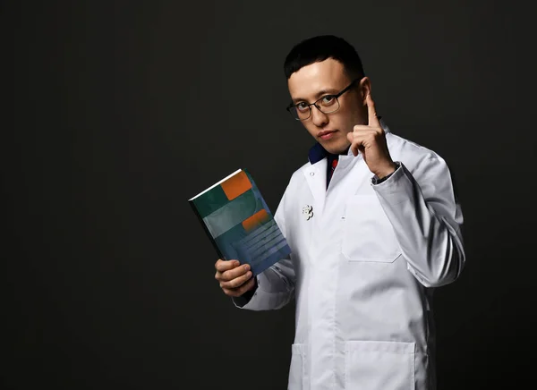 Young man doctors in white medical uniform gown stands holding professional books with blank cover in hand and finger up