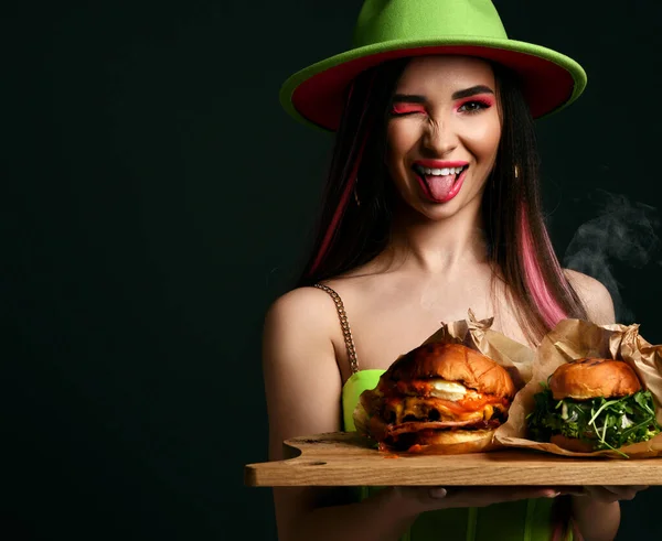 Mooie vrouw houden twee ambachtelijke grote hamburger sandwiches vreugdevol steekt uit tong en knipogen met stoom rook in groene hoed — Stockfoto