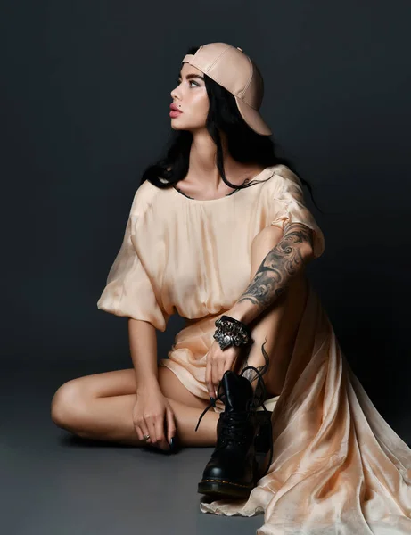 Jeune femme brune sexy avec tatouage sur le bras et perçage sur le visage en robe et bottes massives assis sur le sol et regardant de côté — Photo
