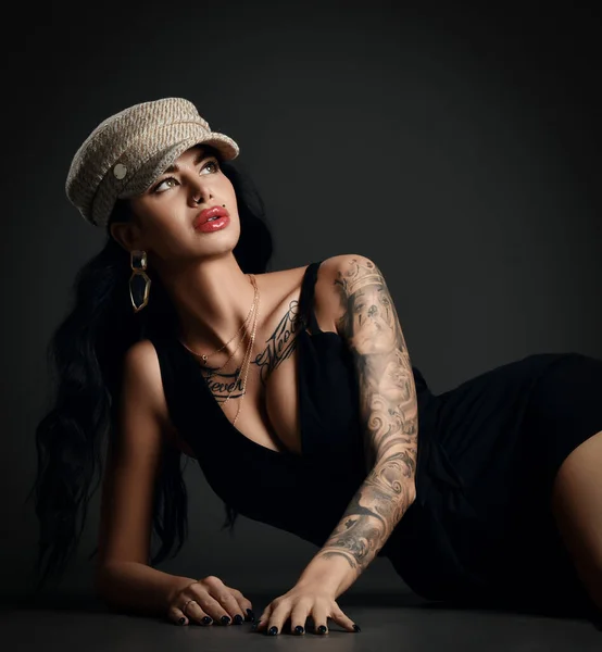 Mulher sexy com lábios pouty e tatuagem em vestido de noite preto com decote profundo está deitado no chão olhando canto superior — Fotografia de Stock