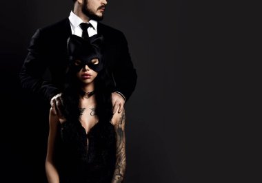 Siyah iç çamaşırlı ve seksi maskeli esmer kadın takım elbiseli adamla oturuyor ve ona arkadan sarılıyor.