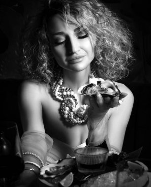 Güzel kıvırcık saçlı bir kadının deniz ürünleri restoranında istiridye yerken ve koyu renk şarap içerken siyah beyaz görüntüsü.