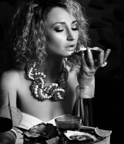 Imagen en blanco y negro de una hermosa mujer rizada en un restaurante de mariscos comiendo ostras y bebiendo vino en la oscuridad — Foto de Stock