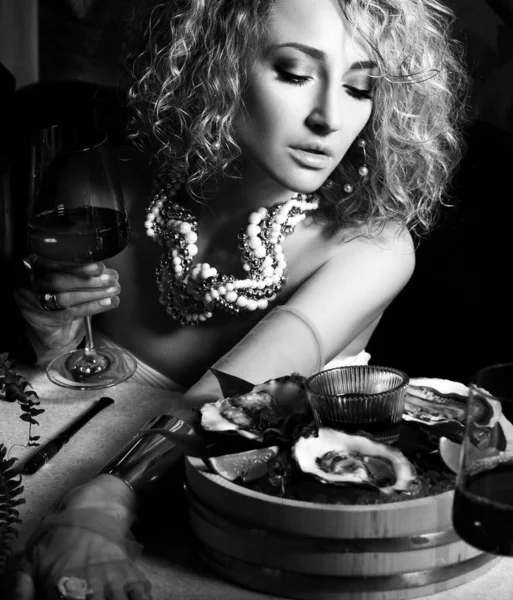 Imagen en blanco y negro de una hermosa mujer rizada en un restaurante de mariscos comiendo ostras y bebiendo vino en la oscuridad — Foto de Stock