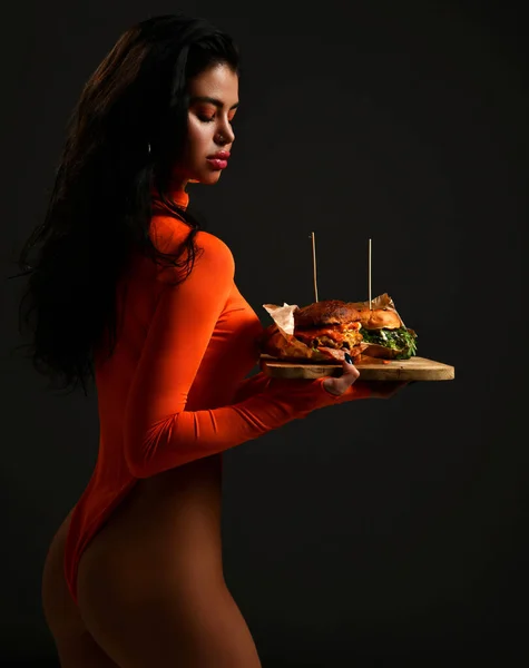 Mulher bonita morena em vermelho sexy bodysuit detém carrega dois artesanato grandes sanduíches de hambúrguer com fumaça de vapor na bandeja de madeira — Fotografia de Stock