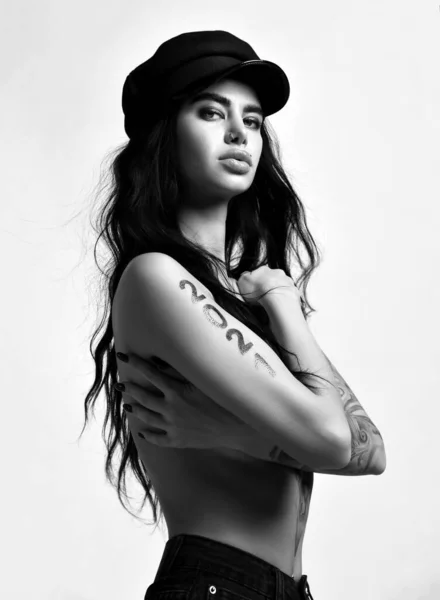 Portrait noir et blanc de femme brune nue avec des lèvres pouty percing et tatouage 2021 sur les bras debout latéralement — Photo