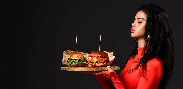 Wanita cantik berambut cokelat dengan mata tertutup dan bibir cemberut dengan bodysuit merah memegang dua burger besar segar di baki kayu. Banner — Stok Foto