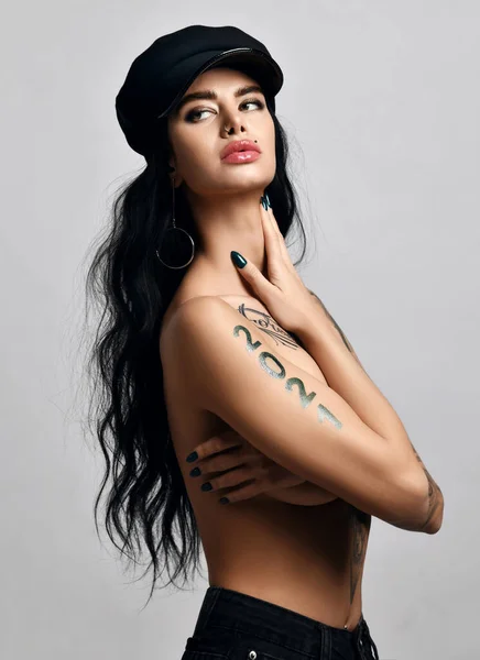 Portrait de femme brune nue avec des lèvres pouty percing et tatouage 2021 sur les bras debout sur le côté et regardant en arrière — Photo