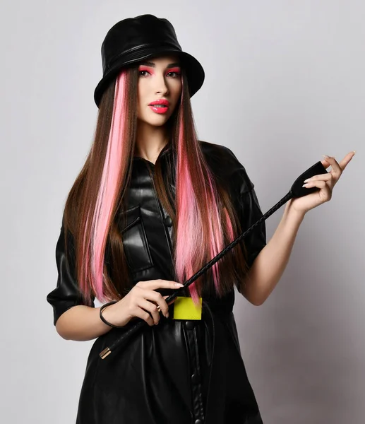 Nő színes haj és duzzadt ajkak visel fekete bőr ruha és kalap állványok gazdaság lovaglás termés ostor a kezében — Stock Fotó