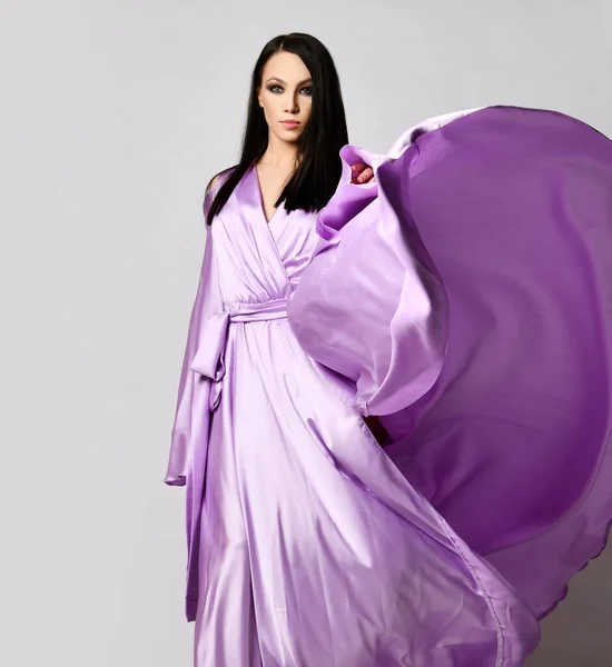 Porträt einer anmutigen brünetten Frau in fliederfarbenem Maxi-Abendkleid Seidenkleid steht mit im Wind flatterndem Saum — Stockfoto