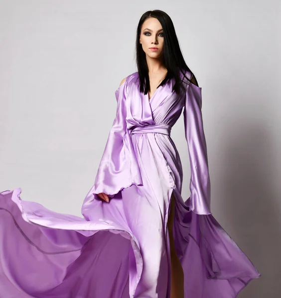 Junge anmutige hübsche brünette Frau in lila langem Maxi-Kleid Seidenkleid steht und hält Saum flatternd im Windsaum — Stockfoto