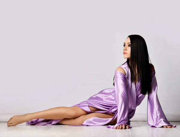 Anmutige sinnliche brünette Frau im lila Maxi-Kleid Seidenkleid liegt mit nackten Beinen auf dem Boden und schaut zur Seite — Stockfoto