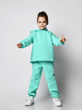 Frolic boy in modern green, mint color hoodie, pants and white sneakers stands holding hands up pointing with fingers