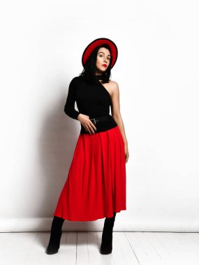 Şık siyah kolsuz bluzlu, kırmızı etekli ve şapkalı güzel esmer kadın kameraya bakıyor.