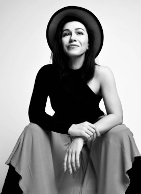 Siyah beyaz portre, yetişkin güzel esmer kadın modern siyah kolsuz bluz, maksi etek ve şapka içinde oturuyor.