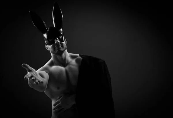Naked topless muscular man in black rabbit mask and with jacket on shoulder stands with his arm outstretched to camera