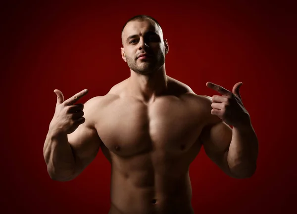 Strong muscular unshaved brutal man, bodybuilder standing with naked chest and pointing fingers at himself