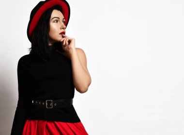 Siyah kolsuz bluz, kırmızı etek ve kırmızı-siyah geniş kenarlı şapka giyen seksi yetişkin kadın düşünceli bir şekilde yana bakıyor.