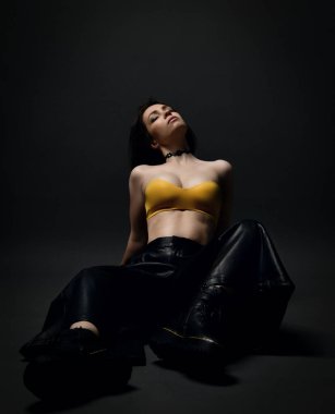 Deri pantolonlu, genç, seksi, uyuşuk esmer kadın. Acımasız Choes ve sutyen. Kafası geriye atılmış şekilde yerde oturuyor.