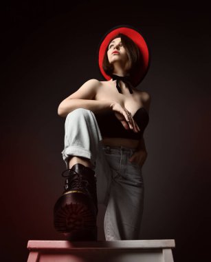 Kendine güveni tam seksi bir kadın, iri çizmeli, siyah sütyenli, kot pantolonlu ve geniş kenarlı şapkalı sandalyede oturuyor.