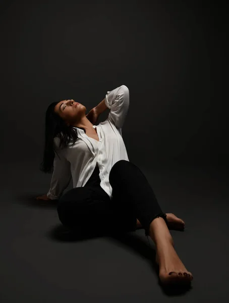 Adult brunette woman in official suit white blouse and black pants sits barefooted on floor with her head thrown back